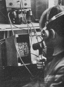 A man in headphones holding a close mike looks at a television monitor