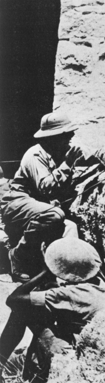 Two men crouch behind a stone wall